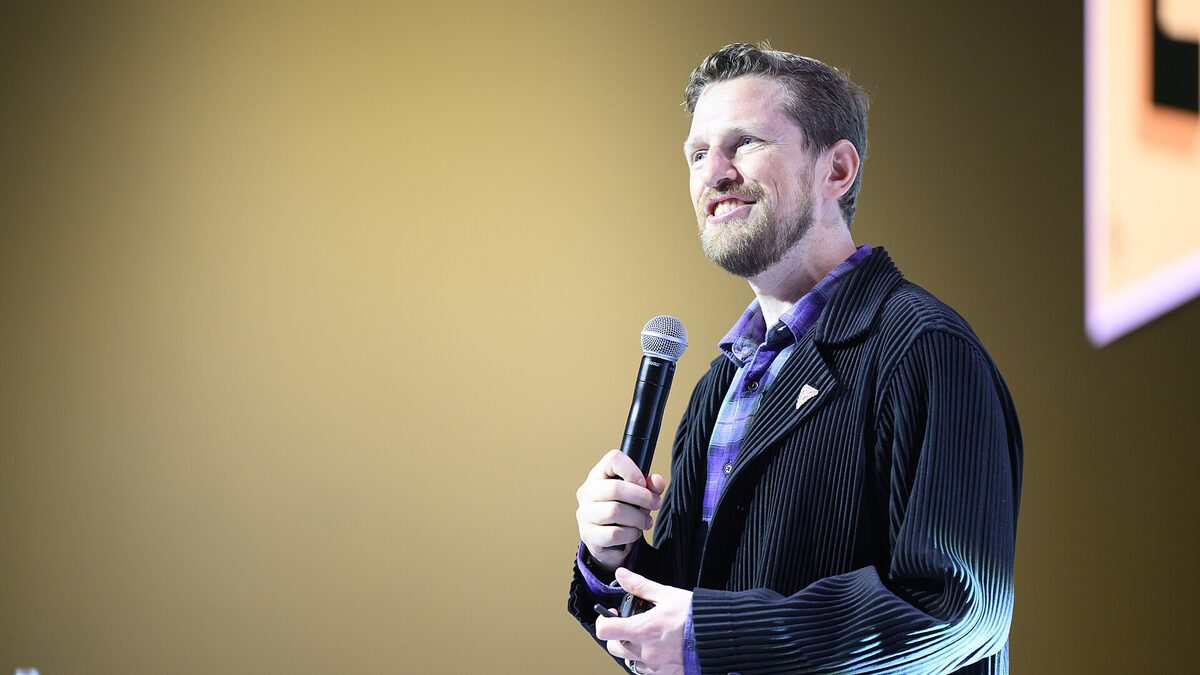 Matt Mullenweg speaking on stage with a handheld microphone against a plain gold backdrop, wearing a dark pleated jacket over a purple plaid shirt.