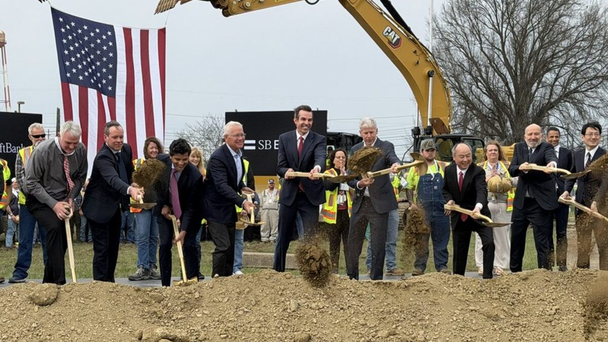 Officials in suits and safety vests push ceremonial shovels into a mound of dirt at the Piketon, Ohio groundbreaking, with a large US flag, an SB Energy banner and a Caterpillar excavator behind them.