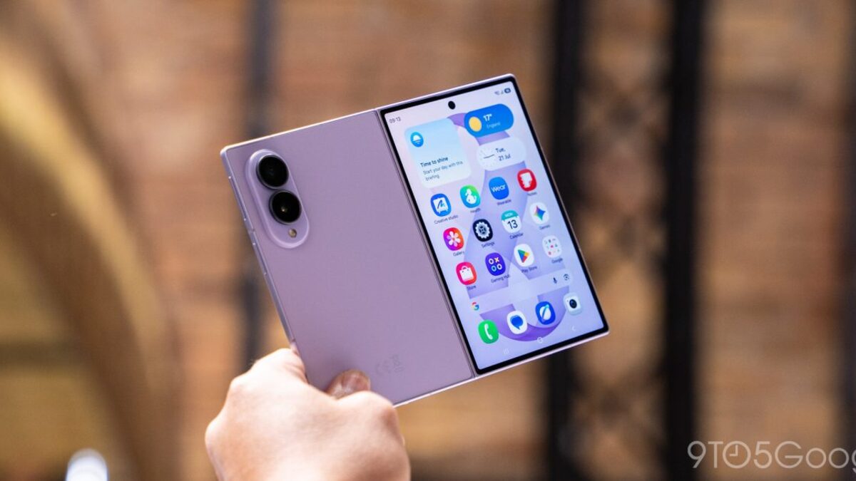 A hand holds a lavender Galaxy Z Fold 8 partly folded outdoors, showing the rear camera bump on one half and the inner display's home screen on the other