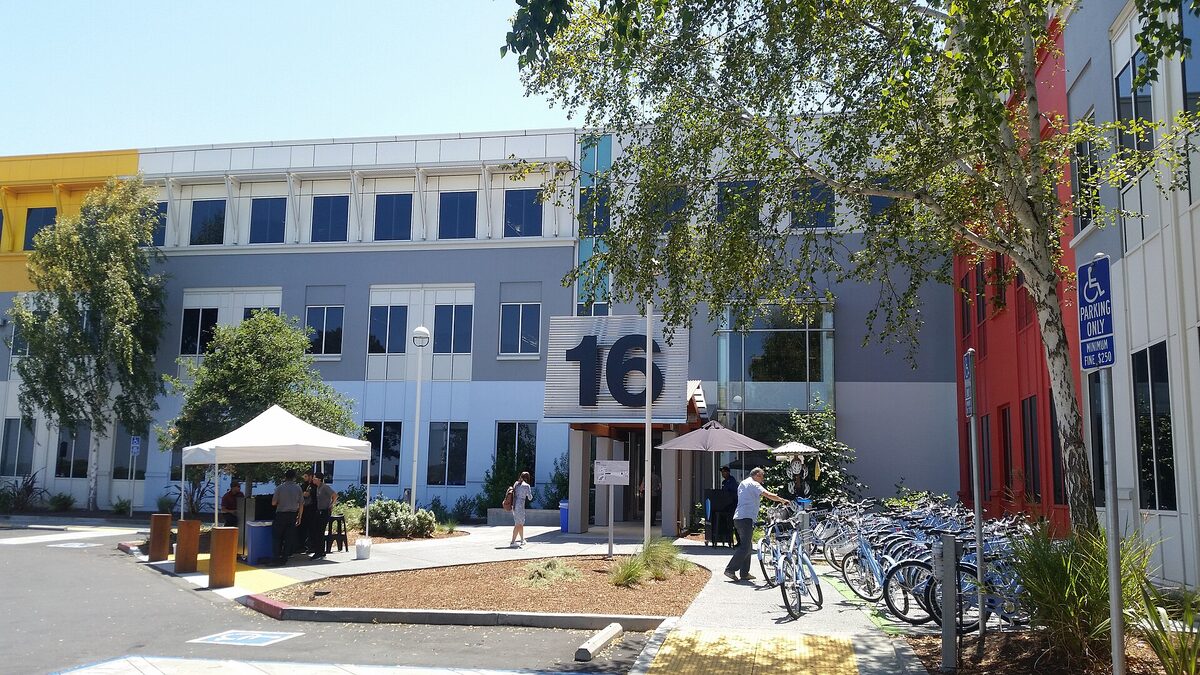 A low-rise office building marked with a large numeral 16 on Meta's Menlo Park campus, with staff, sun umbrellas and a rack of blue bikes outside