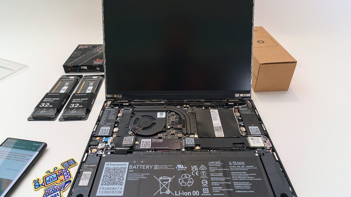 An open Framework Laptop 13 in DIY configuration on a white desk, mainboard exposed, with two 32GB memory modules and an SSD beside it