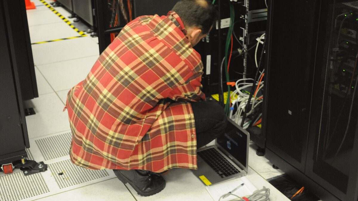 A technician crouches with a laptop to service a black server rack in a data center, colored network cables running down the side