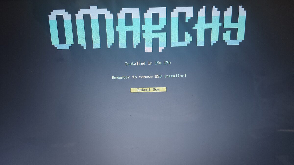 A photo of a monitor showing Omarchy's installer completion screen: a pixel-art OMARCHY wordmark above the line Installed in 19m 17s and a Reboot Now button.